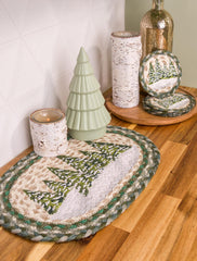 Winter place mat with green pine tree design and white snow on wood counter with birch tree candle holders. 