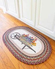 Braided rug with wheelbarrow full of flowers design and jute in rust, yellow, grey, and maroon colors.